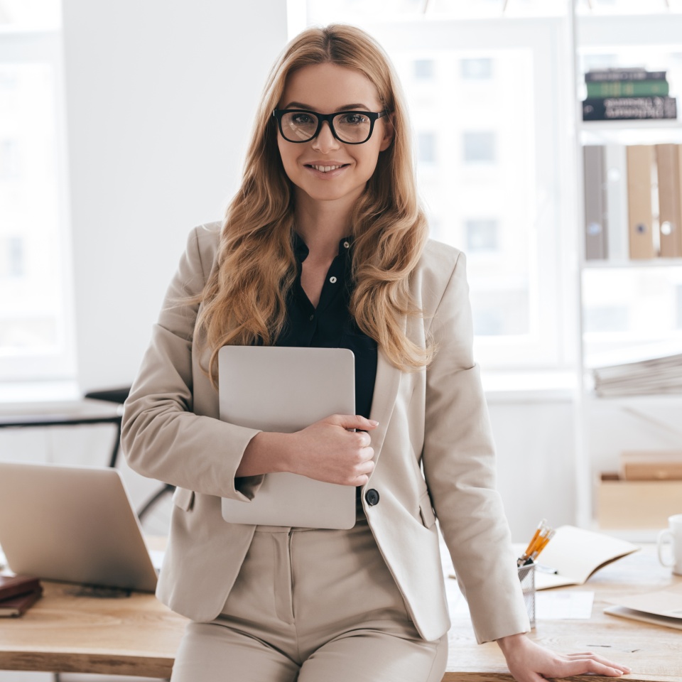 Confident business expert. Attractive young smiling woman in smart casual wear holding digital tablet and looking at camera while leaning on desk in creative office