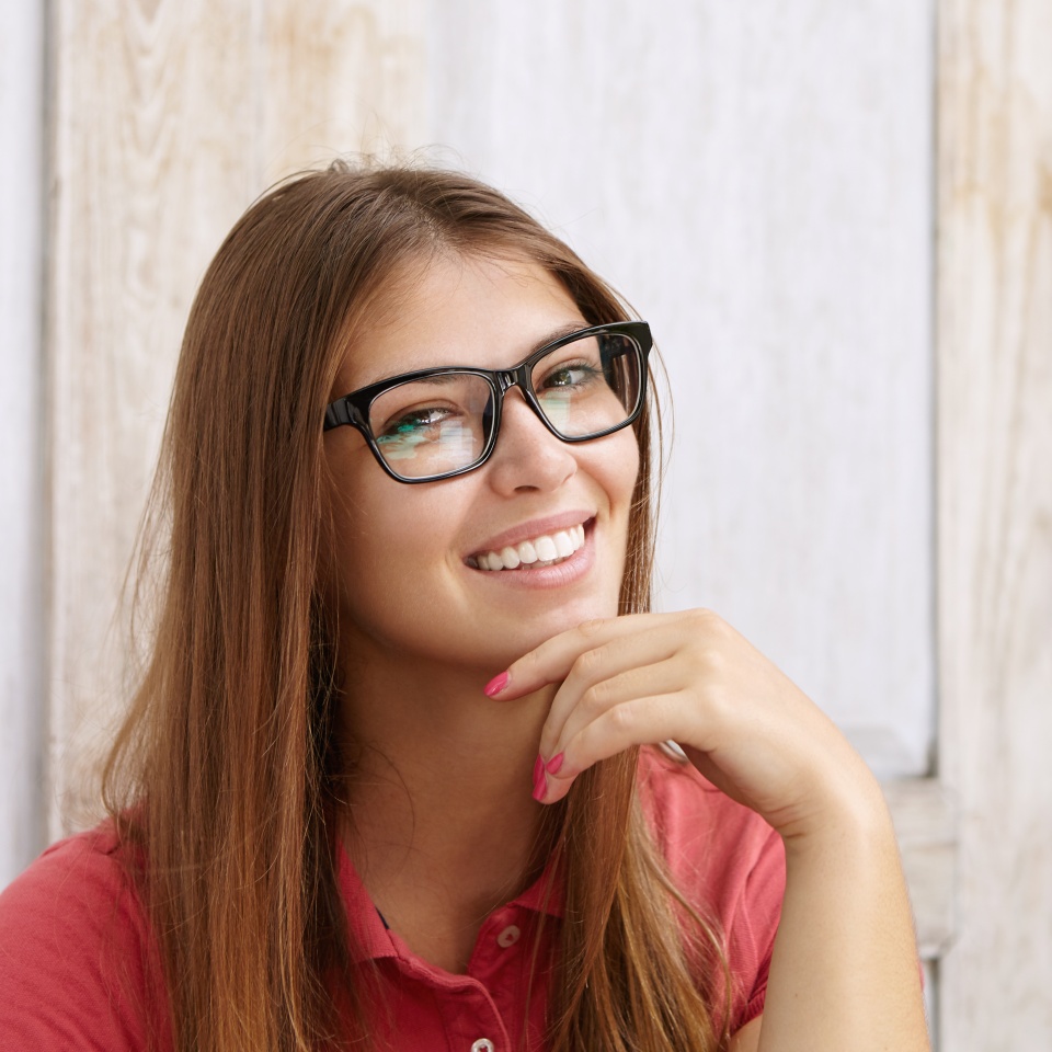 Cheerful attractive Caucasian girl in rectangular glasses posing indoors against wooden wall background with copy space for your text or advertising content holding hand on her chin and smiling