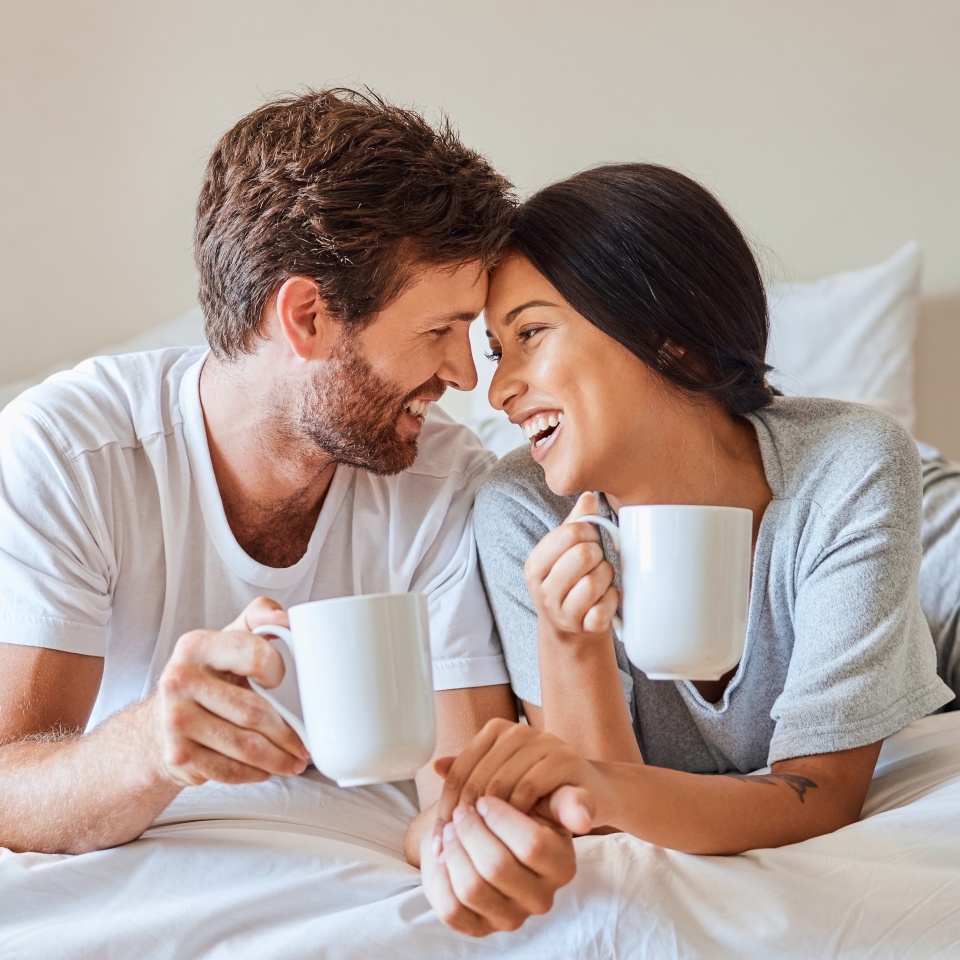 Love, happy and a couple with coffee in bed on beautiful morning at home. Weekend, wake up and smile, woman and man relax with hot drink in bedroom. Romance, happiness and drinking sweet tea together