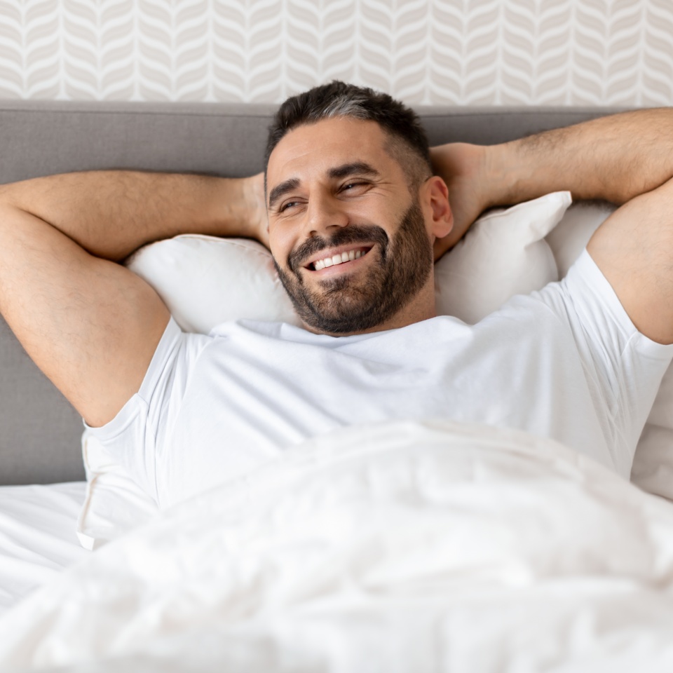 Bearded middle aged man smiling as he lying in bed holding hands behind head, enjoying morning after good night sleep in cozy bedroom at home. Health, relaxation and comfort